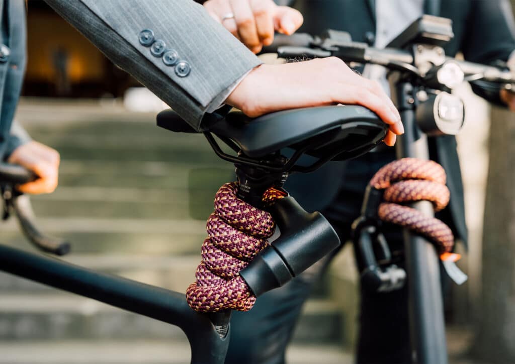Model in een grijs colbert houdt fiets vast bij het zadel, met de tex-lock orbit in chateau red flexibel om de stuurstang gewikkeld